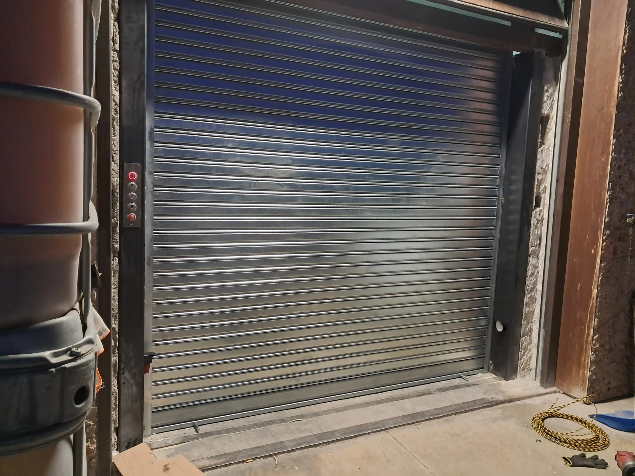 Persiana metálica enrollable cerrada junto a panel de control con botón rojo iluminado.