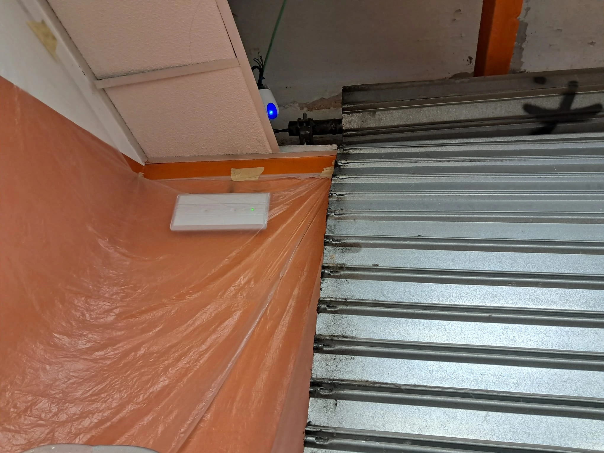 Parte superior de una persiana metálica junto a una pared naranja con un dispositivo electrónico.