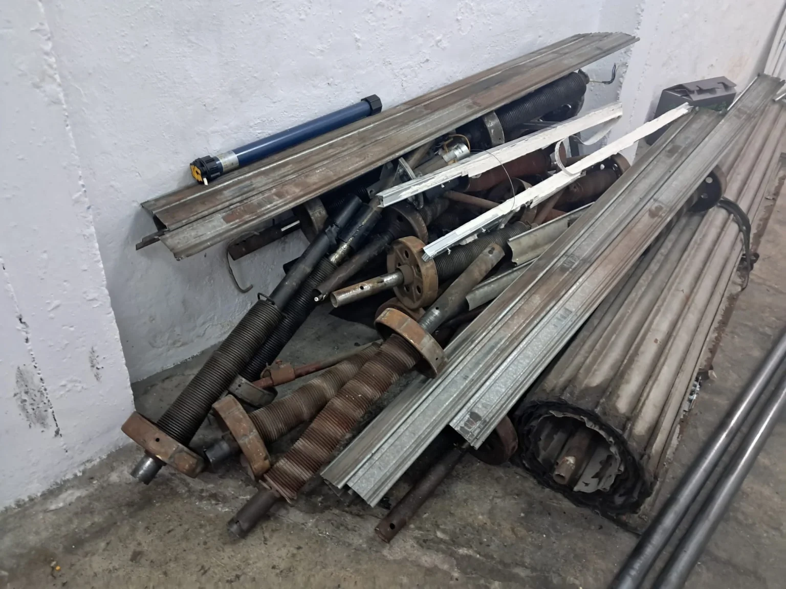Montón de piezas de persiana enrollable desmontadas (muelles, ejes, tambores y motor) con óxido.
