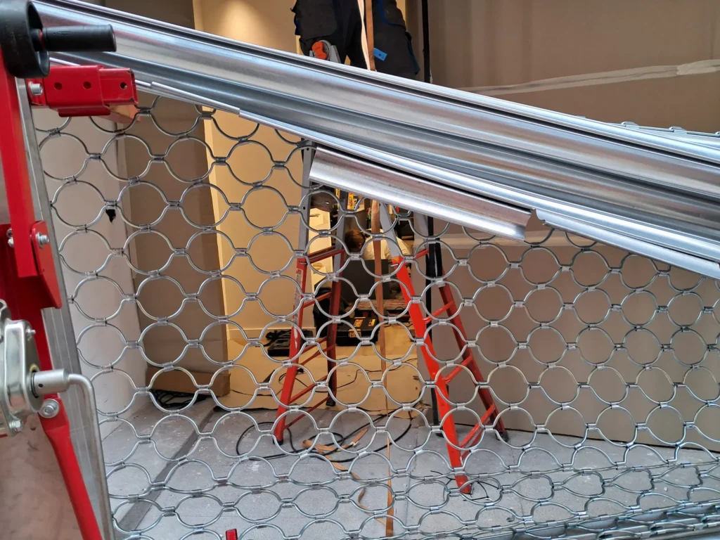 mantenimiento de persianas metalicas - Persiana metálica de panal parcialmente instalada y doblada, con escalera roja y operario trabajando detrás.