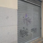 Persiana metálica cerrada con grafiti, fachada de piedra y hojas secas en el suelo.