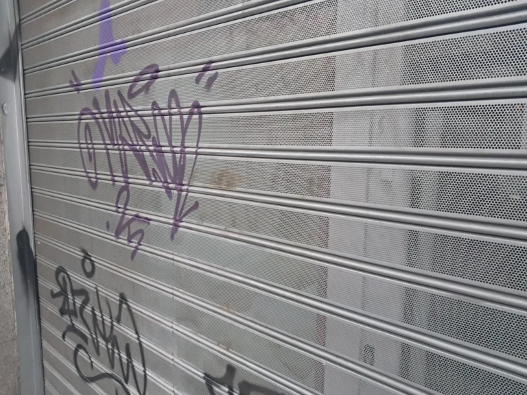 Persiana metálica perforada en primer plano, cubierta de grafiti en morado y negro.