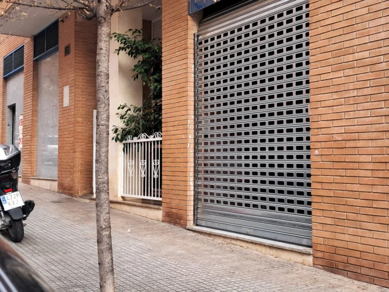 Persiana metálica cerrada con grafiti sobre fachada de ladrillo; scooter aparcado y árbol en la acera