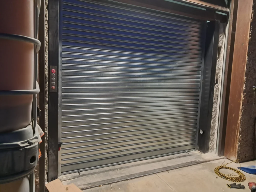Persiana metálica enrollable cerrada junto a panel de control con botón rojo iluminado; en primer plano cuerda amarilla y negra enroscada, guantes y piezas de taller sobre suelo de hormigón.