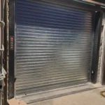 contacto - Persiana metálica enrollable cerrada junto a panel de control con botón rojo iluminado; en primer plano cuerda amarilla y negra enroscada, guantes y piezas de taller sobre suelo de hormigón.