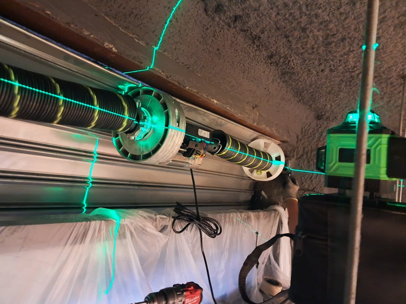 Instalación de mecanismo de puerta de garaje con láser verde cruzando el eje del muelle y tambor; cable y equipo cerca.
