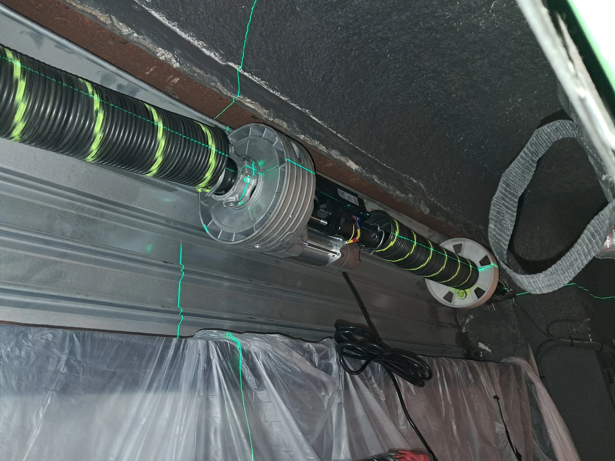 persianasmetalicas75 Mecanismo de motor y muelle de puerta de garaje con líneas láser verdes de alineación; cable eléctrico y protección plástica abajo.