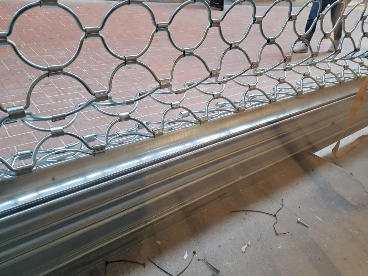 Reja de seguridad metálica enrollable parcialmente instalada con patrón de semicírculos; marco horizontal inferior y suelo de ladrillo visible detrás