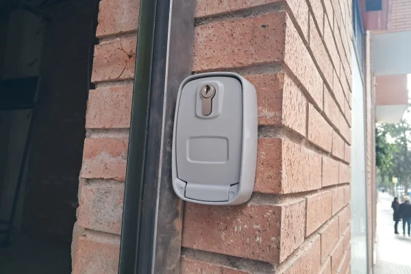 Caja de seguridad metálica con cerradura para desbloqueo de motor de persiana, instalada en pared de ladrillo.