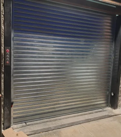 Persiana metálica enrollable cerrada junto a panel de control con botón rojo iluminado.
