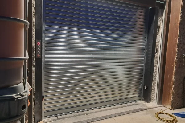 Persiana metálica enrollable cerrada junto a panel de control con botón rojo iluminado.