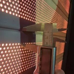 Calibrador digital midiendo el espesor de un panel metálico perforado, con iluminación trasera.