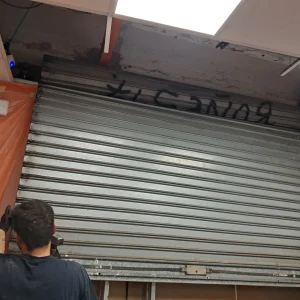Instalador trabajando en una persiana metálica enrollable bajo techo interior con paneles desmontados.